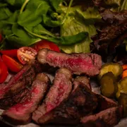 Porções: Picanha Grill & Salada - Picanha nobre grelhada na brasa aproximadamente 200g in natura, fatiada em tiras suculentas ao ponto da casa. Acompanha mix de folhas frescas, picles, tomatinhos e nossa farofa artesanal.